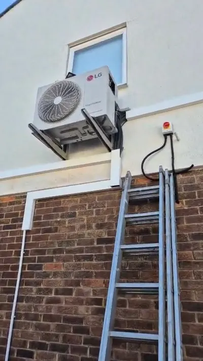 Outdoor air conditioning unit on wall brackets above brick, with isolator switch and pipework in trunking