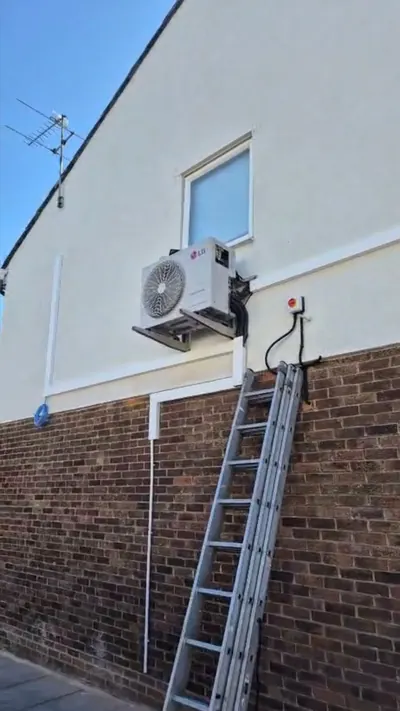 Outdoor unit mounted high on a rendered wall with a ladder alongside